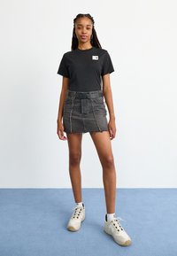 Tricou negru din bumbac cu logo, fustă mini din denim gri cu margine destrămată și cusături contrastante, purtată cu adidași deschisi pe un podea albastră.