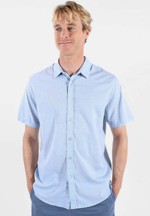 Homme blond portant une chemise bleue claire à manches courtes boutonnée et un pantalon bleu, les mains dans les poches, se tenant devant un fond blanc.