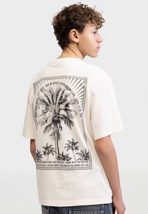 Νεαρός με σγουρά μαλλιά που φοράει λευκό T-shirt με μαύρες εικόνες φοίνικα και εμψυχωτικό κείμενο στην πλάτη.