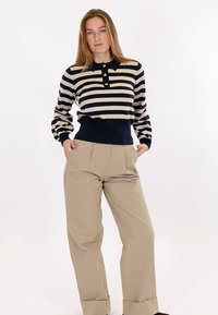 Donna in piedi con le mani in tasca, che indossa una maglia a maniche lunghe a righe blu navy e beige e pantaloni beige a gamba larga, su sfondo bianco.