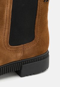 Botte en daim marron avec un panneau latéral élastique noir et une semelle épaisse en caoutchouc noir avec une semelle extérieure texturée, gros plan sur la zone du talon.