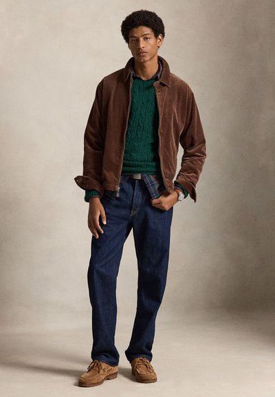 Joven con chaqueta de pana marrón, suéter verde de punto con trenzas sobre camisa de cuadros, vaqueros azul oscuro y zapatos de ante marrón.