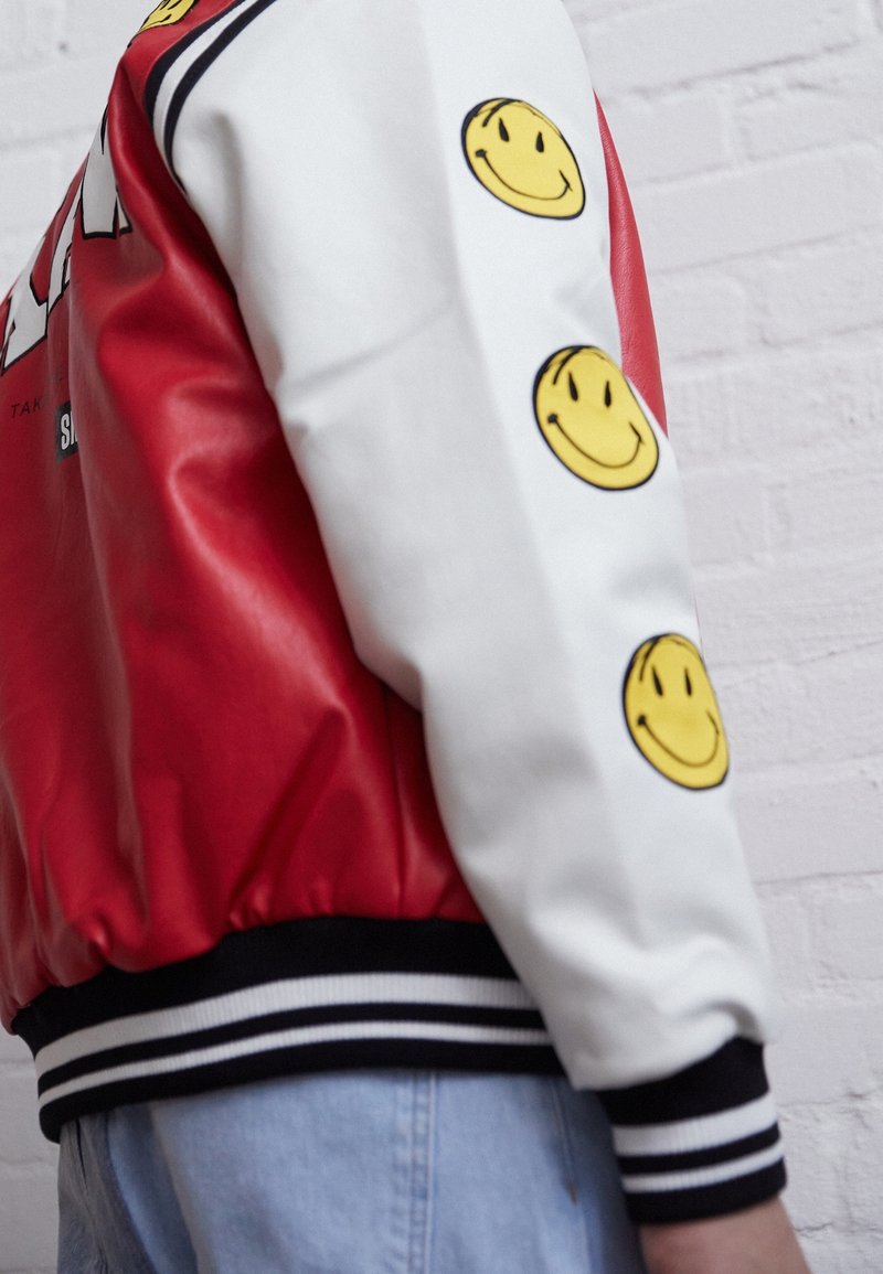 Röd och vit bomberjacka med gula smiley-ansiktsgrafik på ärmarna, ribbade ärmslut och nederkant, med en glänsande yta och djärv design.