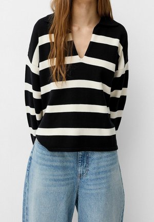 Femme portant un pull à col en V à larges rayures horizontales noires et blanches et un jean taille haute bleu clair, debout devant un fond blanc.