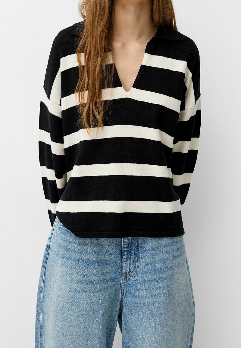 Femme portant un pull à col en V à larges rayures horizontales noires et blanches et un jean taille haute bleu clair, debout devant un fond blanc.