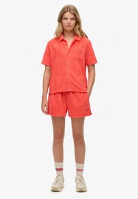 Ensemble en tissu éponge corail composé d'une chemise et d'un short avec un motif de feuille texturé. La chemise est dotée d'un col et d'une poche ; associé à des chaussettes jusqu'aux genoux et des baskets.