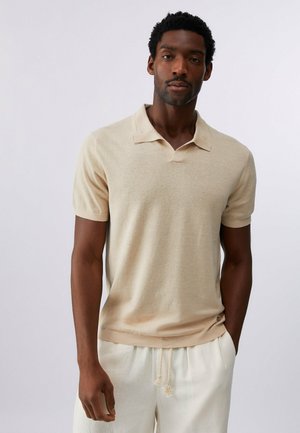 Uomo che indossa una polo beige a maniche corte e pantaloni chiari con coulisse, in piedi davanti a uno sfondo chiaro e uniforme.