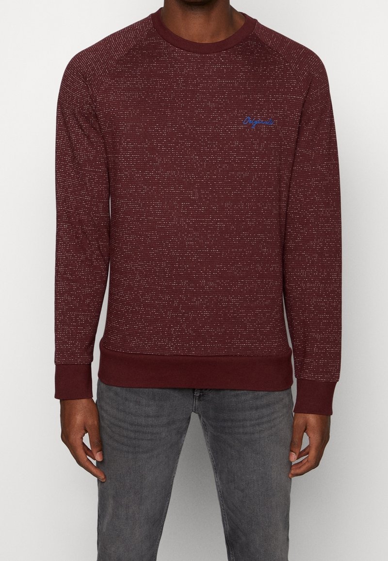 Homme portant un pull col rond lie de vin à motifs avec un texte bleu "Originals" sur la poitrine et un jean gris sur un fond clair.