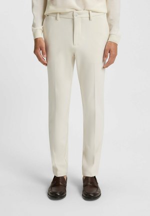 Beige dressbukser med lige ben-design, der har flad front, sidelommer og knaplukning, parret med mørkebrune sko.