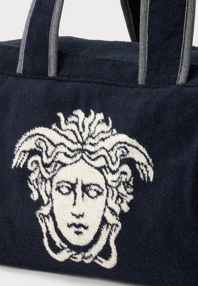 Τσάντα από υφασμάτινο σκούρο μπλε ύφασμα με μαύρες και άσπρες ριγέ λαβές, με λευκό κεντητό σχέδιο κεφαλής Μέδουσας στο εμπρός μέρος.