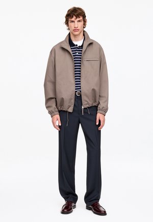 Jeune homme debout portant une veste beige zippée, un polo rayé bleu marine, un pantalon foncé et des chaussures habillées bordeaux sur fond blanc.