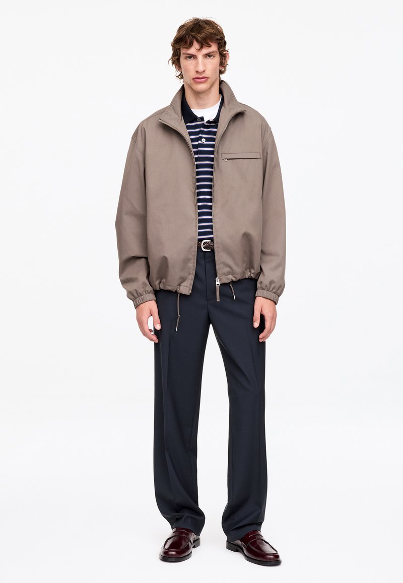 Jeune homme debout portant une veste beige zippée, un polo rayé bleu marine, un pantalon foncé et des chaussures habillées bordeaux sur fond blanc.