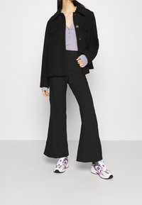 Černá bunda s předními kapsami přes světle fialové tričko, v kombinaci s černými zvonovými kalhotami a barevnými sportovními botami. Hladké, strukturované materiály.