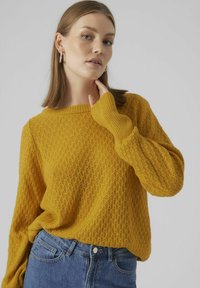 Senapsgul stickad tröja med en strukturerad diamantdesign, bred båt-urringning och ribbade ärmar, i kombination med högmidjade blå jeans.