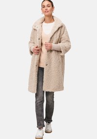 Beige nepbontjas met een brede kraag en drukknoopsluitingen, gedragen over een licht truitje, gecombineerd met grijze spijkerbroek en witte sneakers.