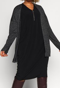 Abito nero con scollatura a V e cerniera argentata, sovrapposto a un cardigan grigio scuro con maniche aperte. Tessuto morbido, vestibilità comoda.