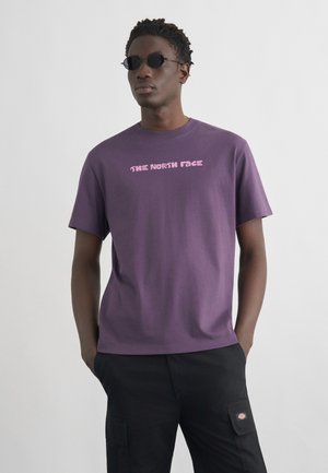 Man wearing purple The North Face t-shirt and black cargo pants, standing with hands in pockets against plain background.