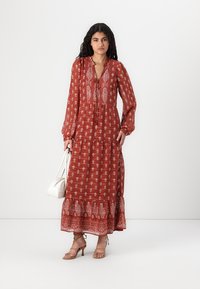 Robe maxi en rouille avec des motifs floraux blancs, manches longues, décolleté en V avec des liens, et un ourlet à volants. Accentuée par un sac à main blanc.