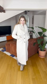 Pikk valge mantel karusnahkrauaikraega, millel on viis nööpi, kaks küljetaskut ja sirge lõige, kantud helehallide pükste ja tossude kohal.