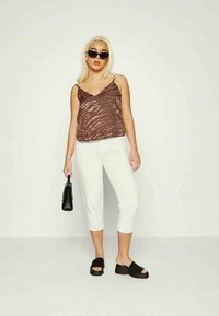 Top de alças castanho com padrão, calças brancas curtas, sandálias pretas de plataforma e uma pequena bolsa preta. Modelo posando contra um fundo liso.