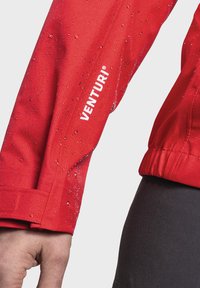 Red waterproof jacket with textured fabric, visible water droplets, and white "VENTURI" logo on the sleeve. Adjustable cuffs at the wrist.