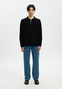 Maglione a maglia navy con collo a zip, abbinato a jeans a gamba dritta azzurri chiari e scarpe in pelle nere. Design semplice e texture liscia.