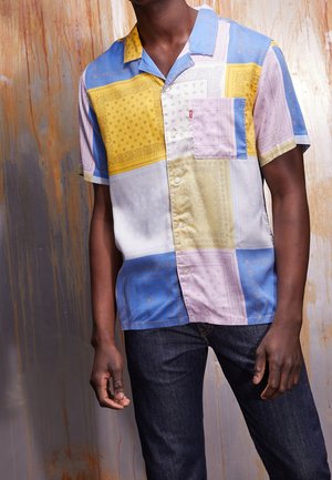 Homme portant une chemise à manches courtes à boutons avec un motif patchwork pastel multicolore et un jean bleu foncé, debout devant un mur texturé.