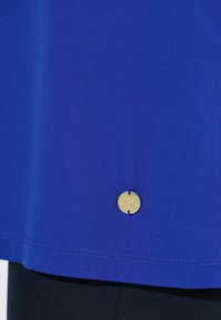 Niebieska tkanina z widocznym przeszyciem na brzegu, posiadająca małą, okrągłą złotą metalową plakietkę blisko dolnej krawędzi z wytłoczonym napisem.