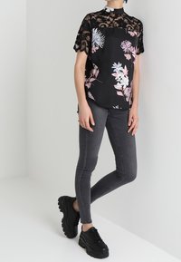 Blusa negra con flores y mangas cortas de encaje, con patrones de flores en rosa y azul. Combinada con jeans ajustados grises y zapatillas negras gruesas.