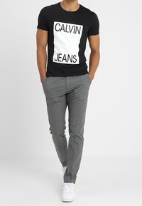 Zwarte katoenen t-shirt met een witte rechthoekige grafiek met de woorden "CALVIN" en "JEANS." Gecombineerd met grijze slim-fit broek en witte sneakers.