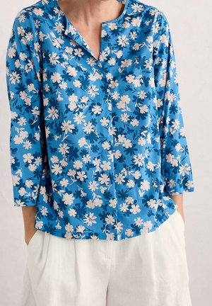 Blå blomsterbluse med hvide og mørkeblå blomster, med rund halsudskæring og 3/4-ærmer, båret med hvide bukser.