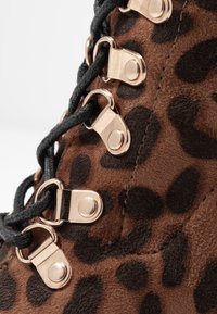 Botte en daim marron avec imprimé léopard noir, lacets noirs passant à travers des œillets et crochets en métal doré.