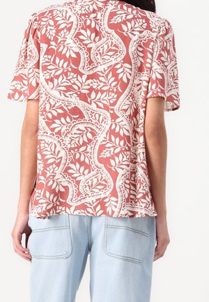 Personne portant une chemise rouge à manches courtes avec des motifs feuillus blancs et un jean bleu clair, vue de dos.