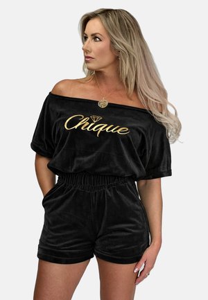 Kobieta w czarnym welurowym kombinezonie z odkrytymi ramionami i złotym napisem "Chique", z rękami w kieszeniach, patrząca w lewo na białym tle.