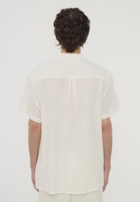 Camicia a maniche corte con colletto, di colore bianco sporco, realizzata in tessuto leggero con una trama testurizzata, dotata di orlo dritto e piega sul retro.