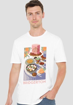 Mann trägt weißes Bridgerton-T-Shirt mit einem bunten Desserttisch, auf dem Kuchen, Macarons, Kirschen und Blumen zu sehen sind.