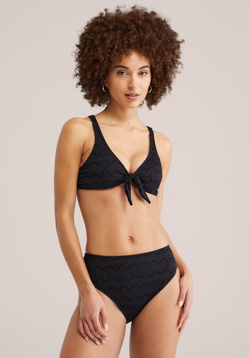 Femme aux cheveux bouclés portant un bikini noir texturé avec un nouage à l'avant, debout devant un fond uni clair, face à l'avant.