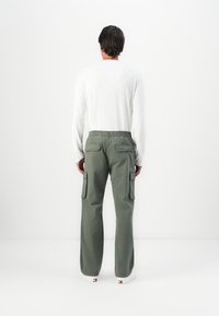 Man wearing white long-sleeve shirt and green cargo pants, standing facing a white wall with hands by his sides.