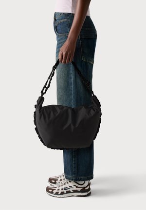 Borsa in tessuto nero con forma morbida e arrotondata, bordi arricciati e dotata di una tracolla spessa e intrecciata. Indossata da una persona con jeans blu.