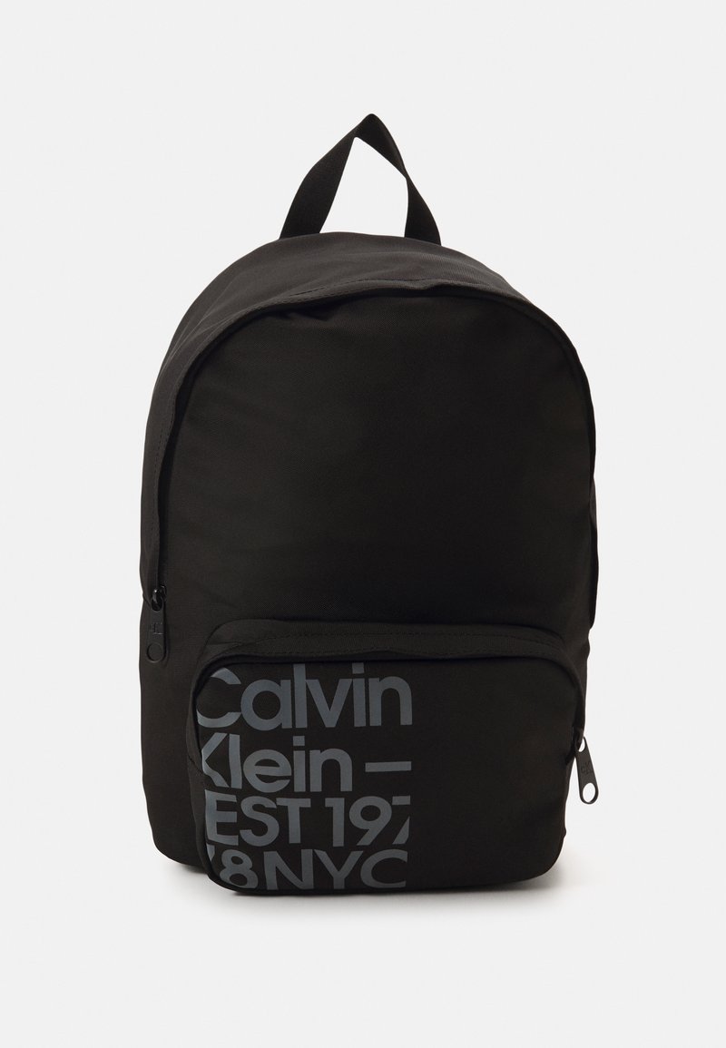 Zaino nero con forma arrotondata, realizzato in tessuto. Presenta una tasca anteriore con zip e testo grigio: "Calvin Klein – EST 1978 NYC."