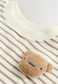 Chemise à manches longues rayée avec un appliqué en forme d'ours en peluche doux et beige près du col, dotée d'un col blanc côtelé et de rayures beiges.