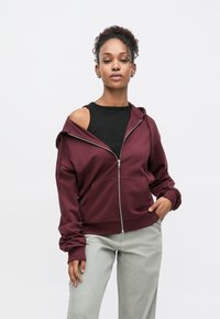 Sweat à capuche zippé bordeaux avec une poche avant, une texture douce, une capuche ajustable et une épaule découverte, associé à un pantalon gris clair.