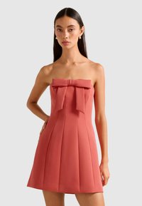 Frau in einem trägerlosen, korallfarbenen Minikleid mit einer großen Schleifendetail am Ausschnitt, die vor einem hellgrauen Hintergrund steht.