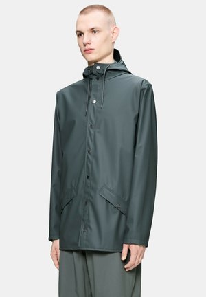 Joven con una chaqueta impermeable con capucha de color gris oscuro, con botones a presión en la parte delantera y bolsillos con solapas inclinadas, de pie frente a un fondo liso.