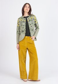 Gilet en tricot vert avec broderies florales, chemise noire et pantalons larges en velours côtelé jaune. Le modèle se tient devant un fond uni.