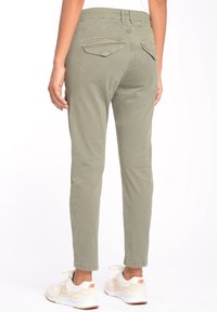 Osoba oblečená v olivově zelených slim-fit kalhotách s kapsami na zadní straně a bílých teniskách s oranžovými logy, stojící na bílém pozadí.