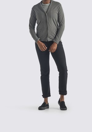 Personne portant une veste grise boutonnée sur une chemise rayée, un pantalon noir retroussé et des chaussures noires sans lacets, debout sur un fond clair.