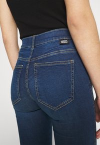 Vaqueros de mezclilla azul oscuro con cintura alta, que cuentan con cinco bolsillos, costuras amarillas contrastantes y una pequeña etiqueta de marca en la parte posterior.