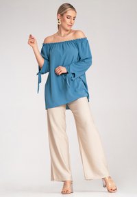 Niebieska bluzka z odkrytymi ramionami, z elastycznym dekoltem, długie rękawy wiązane przy mankietach; zestawiona z beżowymi spodniami o szerokich nogawkach i przezroczystymi sandałami na obcasie.