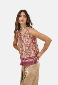 Top sin mangas con estampado floral en rosa y beige, que presenta un lazo en el escote y un fleco en la parte inferior con pompones.